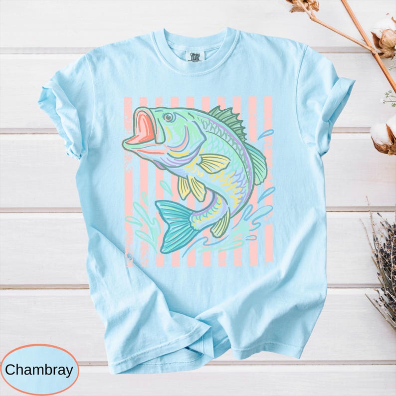 Puede incluir: Camiseta azul claro con una ilustraci&oacute;n colorida de un pez saltando con la boca abierta. El pez est&aacute; representado en tonos de verde, amarillo y azul, sobre un fondo de rayas verticales rosas. La palabra "Chambray" est&aacute; en un c&iacute;rculo en la parte inferior.