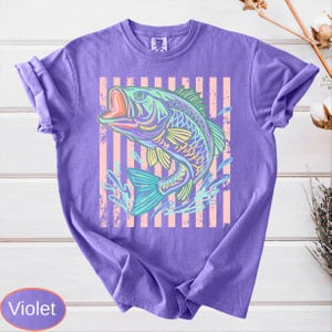 Puede incluir: Camiseta violeta con una colorida ilustraci&oacute;n de un pez con la boca abierta. El pez est&aacute; sobre un fondo de rayas verticales rosas y blancas. La camiseta tiene mangas cortas y cuello redondo.
