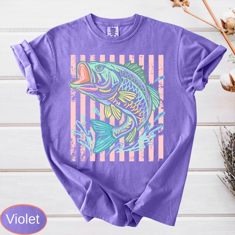 Puede incluir: Camiseta violeta con una colorida ilustraci&oacute;n de un pez con la boca abierta. El pez est&aacute; sobre un fondo de rayas verticales rosas y blancas. La camiseta tiene mangas cortas y cuello redondo.