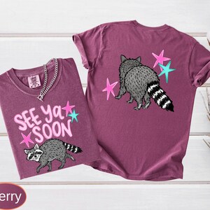 May include: Two berry-colored t-shirts with raccoon graphics. One shirt features the text "SEE YA SOON" with pink and teal stars. The other shirt has a raccoon graphic and stars on the back. Both shirts have short sleeves.