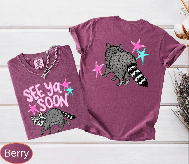 May include: Two berry-colored t-shirts with raccoon graphics. One shirt features the text "SEE YA SOON" with pink and teal stars. The other shirt has a raccoon graphic and stars on the back. Both shirts have short sleeves.