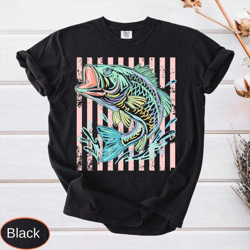 Puede incluir: Camiseta negra con una colorida ilustraci&oacute;n de un pez con la boca abierta, sobre un fondo de rayas rosas y blancas. El dise&ntilde;o del pez incorpora tonos de azul, amarillo y verde, con salpicaduras de agua.