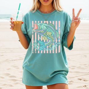 Puede incluir: Camiseta de color turquesa con un gr&aacute;fico de pez colorido, con la boca abierta, sobre un fondo de rayas rosas y blancas. El dise&ntilde;o del pez utiliza tonos azules, amarillos y verdes. La camiseta tiene mangas cortas y un corte holgado.