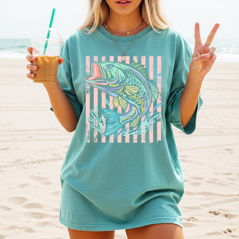 Puede incluir: Camiseta de color turquesa con un gr&aacute;fico de pez colorido, con la boca abierta, sobre un fondo de rayas rosas y blancas. El dise&ntilde;o del pez utiliza tonos azules, amarillos y verdes. La camiseta tiene mangas cortas y un corte holgado.