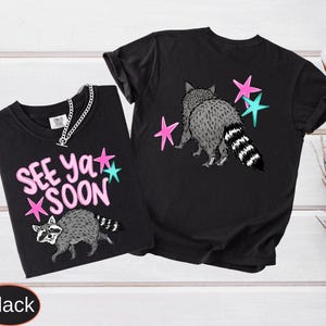 May include: Two black t-shirts with raccoon graphics and the text "SEE YA SOON". The raccoon is gray and white with a striped tail. Pink and teal stars are also featured in the design. One shirt has a silver chain.
