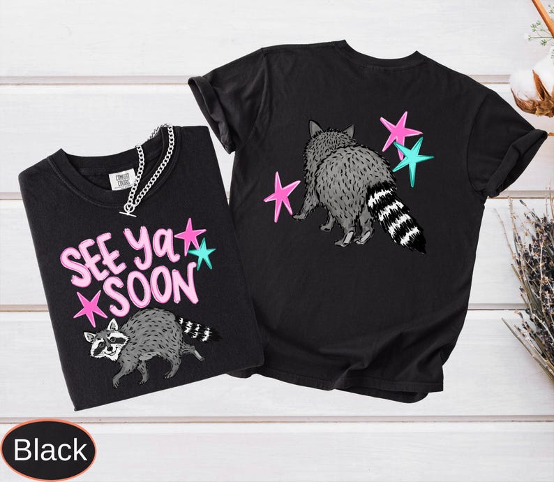 May include: Two black t-shirts with raccoon graphics and the text "SEE YA SOON". The raccoon is gray and white with a striped tail. Pink and teal stars are also featured in the design. One shirt has a silver chain.