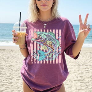 Puede incluir: Una camiseta granate con un gr&aacute;fico colorido de un pez saltando fuera del agua. El pez tiene detalles azules, amarillos y verdes. El dise&ntilde;o est&aacute; sobre un fondo de rayas rosas y blancas. La camiseta es de corte holgado.