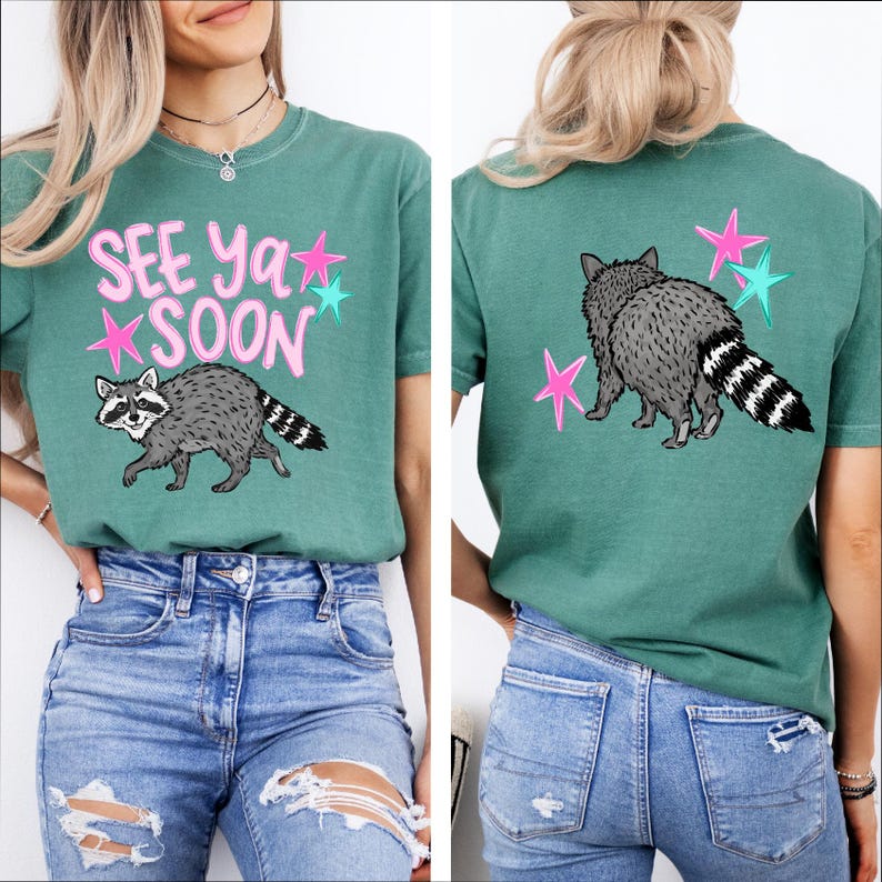 May include: Teal-colored t-shirt with a raccoon graphic and the text "SEE YA SOON" in pink. The raccoon is gray and white with a striped tail. Pink and teal stars surround the text and raccoon.
