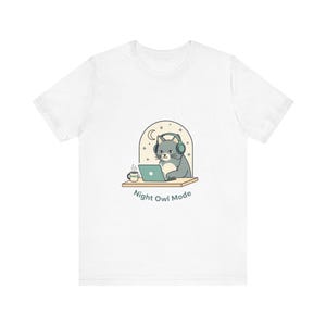 Op de afbeelding: Wit t-shirt met een cartoonkat met een koptelefoon die op een laptop werkt. De kat zit aan een bureau met een kopje koffie, en de tekst "Night Owl Mode" staat eronder.