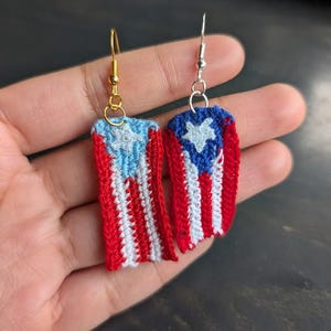 May include: A pair of handmade crochet earrings featuring the Puerto Rican flag design. Each earring displays the flag's red and white stripes, a blue triangle, and a white star. One earring has a gold hook, the other a silver hook.
