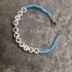 May include: A beaded bracelet featuring a mix of light and medium blue seed beads and white alphabet beads spelling out the words POTS WARRIOR. The bracelet includes a bronze-toned metal lobster claw clasp and a small circular metal jump ring.
