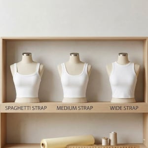 May include: Three white tank tops on mannequins with different strap styles: spaghetti strap, medium strap, and wide strap. The tank tops are displayed on a wooden shelf. Text below each tank top indicates the strap style.
