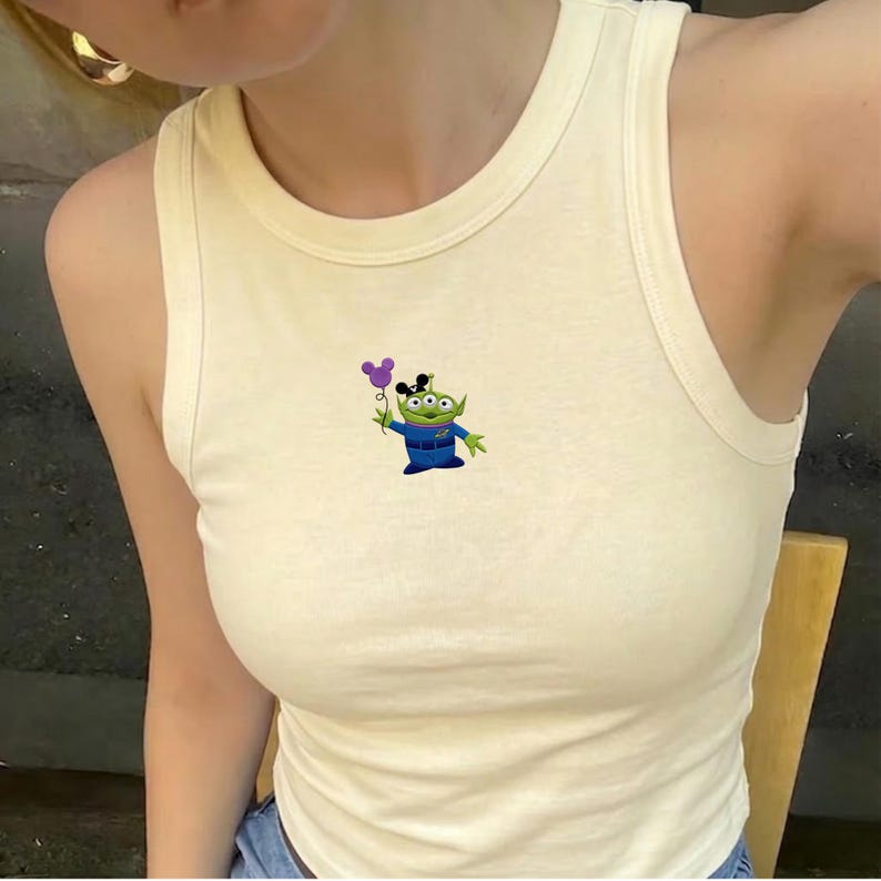 May include: Cream-coloured vest top with a cartoon alien character holding a purple balloon. The alien is green with three eyes and wears a blue uniform. The vest top has a rounded neckline and a sleeveless design.