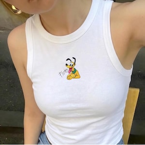 Puede incluir: Camiseta sin mangas blanca con un gráfico de Pluto. El perro de dibujos animados es amarillo con orejas negras y un collar verde. La palabra "Pluto" está escrita en cursiva debajo. La camiseta sin mangas es sin mangas y tiene un cuello redondo.