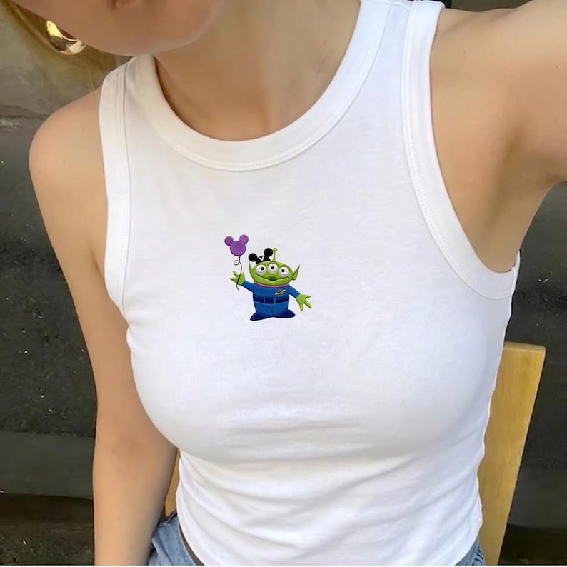 May include: White vest top with a cartoon alien holding a purple Mickey Mouse balloon. The alien is green with three eyes and wears a blue uniform. The vest top is sleeveless and has a rounded neckline.