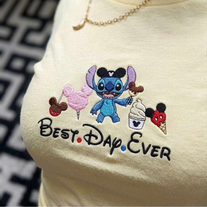 May include: A light yellow t-shirt with an embroidered design featuring the character Stitch, cotton candy, ice cream, and the words "Best. Day. Ever." The design includes Mickey Mouse-themed elements.