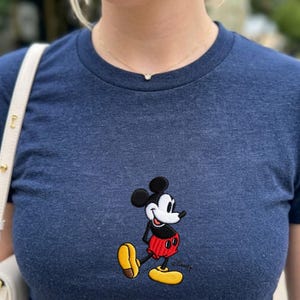 May include: A navy blue t-shirt featuring an embroidered image of Mickey Mouse. The cartoon character is depicted in his classic outfit, with red shorts, yellow shoes, and black gloves. The shirt has a crew neck and short sleeves.