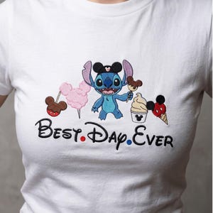 May include: White t-shirt featuring a blue cartoon character with large ears, holding a Mickey Mouse-shaped ice cream. The shirt also has a pink cotton candy, a Mickey Mouse lollipop, and the text "Best. Day. Ever."