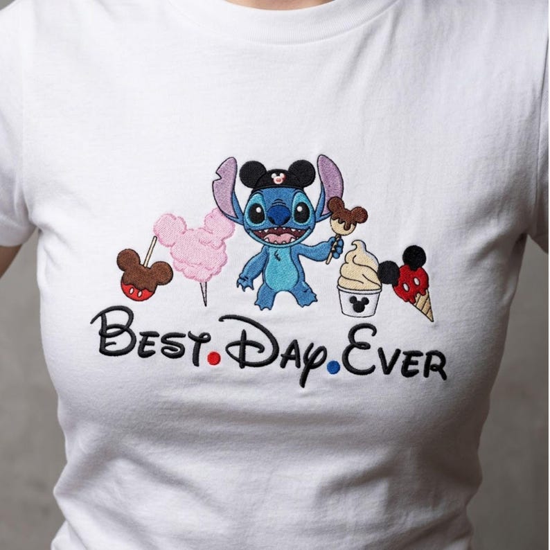 May include: White t-shirt featuring a blue cartoon character with large ears, holding a Mickey Mouse-shaped ice cream. The shirt also has a pink cotton candy, a Mickey Mouse lollipop, and the text "Best. Day. Ever."