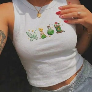 May include: White tank top with embroidered fairy-themed design. The design includes a green butterfly, a fairy, a potion bottle, and a spool of thread with mushrooms. The tank top has a round neckline and is sleeveless.