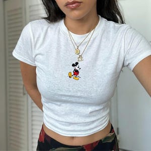 May include: White t-shirt featuring a small Mickey Mouse graphic. The shirt has a crew neck and short sleeves. A gold necklace with two pendants is worn. The shirt is a cropped style.