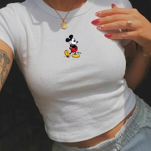 May include: White crop top featuring a classic Mickey Mouse graphic. The cartoon character is depicted in his iconic red shorts, yellow shoes, and black gloves. The shirt has a crew neck and short sleeves, perfect for casual wear.
