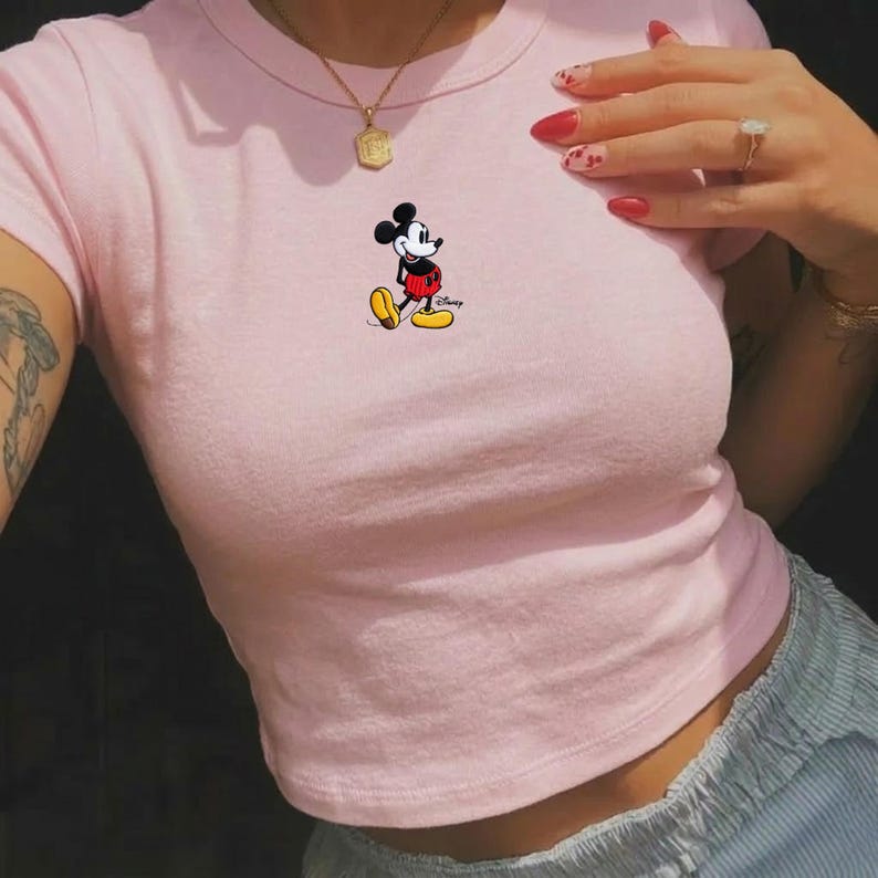 May include: A light pink crop top featuring a small Mickey Mouse graphic. The cartoon character is depicted in his classic outfit with yellow shoes, red shorts, and white gloves. The top is made of a soft material.
