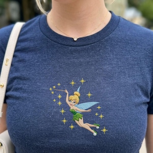 May include: A navy blue t-shirt featuring an embroidered image of a fairy with a wand, surrounded by yellow stars. The fairy is wearing a green dress and blue wings. The shirt is made of a soft, heathered fabric.