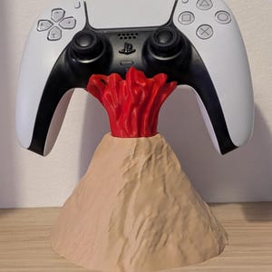 Puede incluir: Un mando blanco y negro de PlayStation 5 sobre un soporte con forma de volcán. El soporte es de color beige con un flujo de lava rojo. El mando tiene una sección central negra con el logotipo de PlayStation.