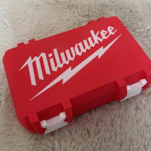 Op de afbeelding: Een rode Milwaukee gereedschapskoffer met een wit logo en bliksemontwerp. De koffer heeft witte sluitingen en is rechthoekig van vorm. De koffer ligt op een lichtgrijze, getextureerde ondergrond.