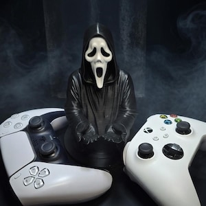 May include: A black and white Scream mask statue with a black hooded robe, flanked by a white PlayStation 5 controller and a white Xbox controller. The controllers have black buttons and joysticks. The background is dark with a smoky effect.