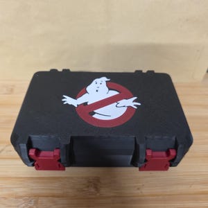 May include: Black rectangular case with red clasps, displaying a white Ghostbusters logo within a red no-ghost emblem. The case rests on a wooden surface.