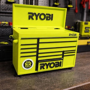 May include: A bright yellow tool chest with black accents and the "RYOBI" logo prominently displayed. The chest has a hinged lid and multiple drawers, with the "RYOBI" logo repeated on the front. The chest is on a brown surface.