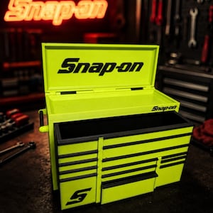 May include: A neon yellow Snap-on tool chest with a black logo on the open lid and front. The metal box features multiple drawers with black trim and a side handle. The Snap-on brand name is printed in black on the lid and the front drawer face.