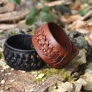 May include: Two leather bracelets, one black and one brown, sit on a natural wood surface. The brown bracelet has a woven design, while the black bracelet has a similar, open pattern. Both are wide cuff style.