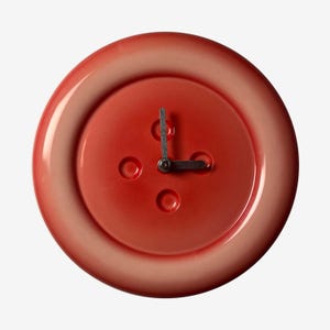 May include: A round, red wall clock designed to resemble a large button. The clock face has three small, circular indentations and black clock hands. The clock's outer edge has a slightly lighter, pinkish hue.
