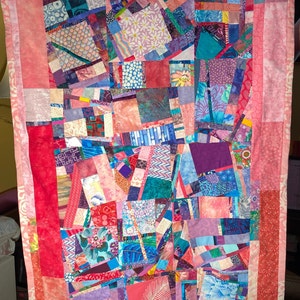 May include: A patchwork quilt with a variety of colorful fabrics, including pink, red, purple, blue, and green. The quilt is made up of squares and rectangles of different sizes and patterns. The quilt is edged with a pink border.
