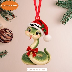 Könnte beinhalten: Ein grüner Schlangen-Anhänger mit einer roten Weihnachtsmütze, auf deren weißem Rand der Name ANNA steht, einer roten Schleife mit kleiner goldener Glocke um den Hals und einem roten Band zum Aufhängen. Die Schlange hat große Augen und eine rote Zunge.