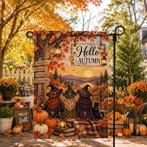 May include: A rectangular garden flag featuring an autumn landscape with three figures in witch hats, pumpkins, and a sign that reads Hello Autumn. The scene includes a tree, lanterns, and a wooden signpost listing Pumpkins, Crisp Air, Cozy Nights, and Brighter Days.