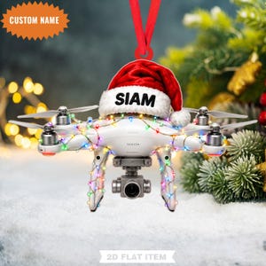 Pode incluir: Um ornamento de drone quadricóptero branco com um gorro de Pai Natal vermelho com o nome SIAM impresso na borda branca. O drone está envolvido por luzes coloridas e pendurado por uma fita vermelha. O texto CUSTOM NAME aparece numa estrela.