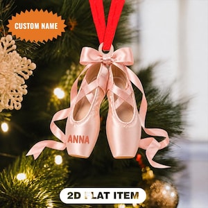Op de afbeelding: Een platte 2D-kerstboomversiering in de vorm van een paar lichtroze balletschoenen met satijnen linten. De linkerschoen is voorzien van de aangepaste naam ANNA in een vetgedrukt lettertype. Een rood lint is bovenaan bevestigd om op te hangen.