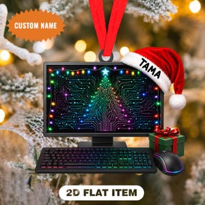 May include: A flat, two-dimensional ornament shaped like a computer monitor with a keyboard and mouse. The screen displays a circuit board pattern in the shape of a Christmas tree with colourful lights. A red Santa hat with the name TAMA is attached.