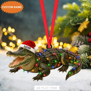 May include: A hanging ornament shaped like an alligator with a textured green and brown body, wearing a red and white Santa hat with the name LUCAS printed on it. The alligator is wrapped in a string of colorful miniature Christmas lights.