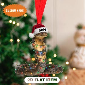 Könnte beinhalten: Ein flacher 2D-Weihnachtsbaumschmuck in Form einer braunen Kobra, die mit einer Lichterkette aus bunten Weihnachtslichtern umwickelt ist. Die Schlange trägt eine rot-weiße Weihnachtsmütze mit dem Namen SAM auf dem Rand und hat ein rotes Band.