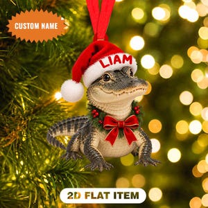 Könnte beinhalten: Ein flacher 2D-Weihnachtsanhänger mit einem Baby-Alligator, der eine rote Weihnachtsmütze mit dem Namen LIAM auf dem weißen Rand trägt. Der Alligator hat einen grünen Kranz um den Hals, der mit einer großen roten Schleife und Beeren verziert ist.