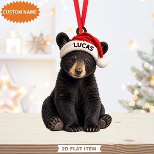 May include: A 2D flat hanging ornament featuring a black bear cub wearing a red and white Santa hat with the name LUCAS printed on the brim. The ornament is suspended by a red ribbon and includes a starburst graphic labelled CUSTOM NAME.