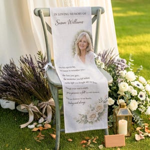 May include: A memorial chair sash with the text "IN LOVING MEMORY OF Susan Williams" and a photo of a woman. The sash has a floral design and a poem. The chair is surrounded by flowers, lavender, and a candle lantern.