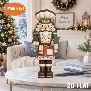 Peut inclure: Une figurine de casse-noisette décorative habillée en chef, avec une toque blanche portant le nom ALEX sur le bord, une veste verte et un tablier rouge. La figurine tient un fouet et un rouleau à pâtisserie, avec une guirlande lumineuse sur le tablier.