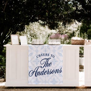 May include: A white bar with a blue and white patterned table runner that reads "Cheers to The Andersons." The bar is set up outdoors with glasses, candles, and a basket. Two framed signs are on the bar.