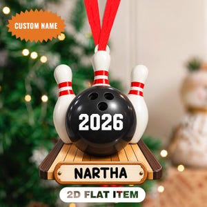 Op de afbeelding: Een platte kerstornament met een bowlingthema, bestaande uit een zwarte bowlingbal met het jaartal 2026 in wit, geflankeerd door drie witte bowlingkegels met rode strepen op een houten baanbasis met de gepersonaliseerde naam NARTHA.